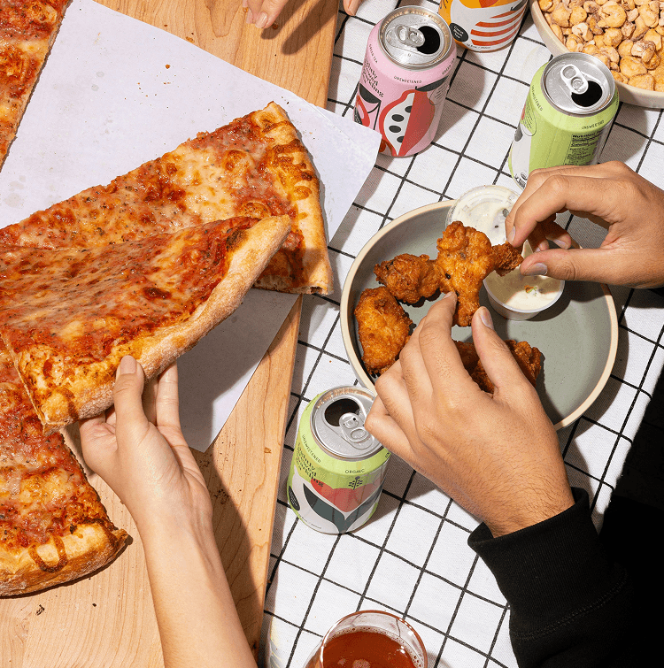 A close-up, overhead shot of people's hands reaching for pizza, fried chicken, and canned drinks on a checkered tablecloth. One hand is lifting a large slice of cheese pizza, while others are dipping pieces of crispy fried chicken into a small bowl of white sauce.