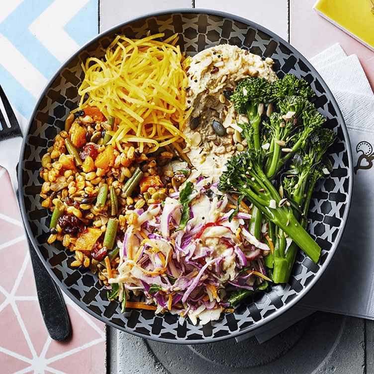 An overhead view of a colorful, patterned bowl filled with a healthy grain and vegetable meal. The bowl contains sections of bright yellow shredded squash or carrots, steamed broccolini, a creamy hummus or dip topped with seeds, and a mix of cooked grains, beans, and sweet potato, next to a shredded cabbage slaw.